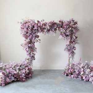 May include: A wedding arch decorated with purple and white artificial flowers. The arch is made of a metal frame and is covered in a variety of flowers, including roses, hydrangeas, and greenery. The arch is standing on a gray concrete floor.