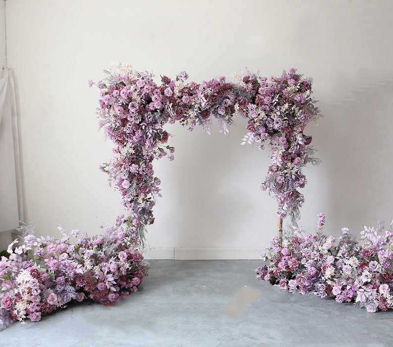 May include: A wedding arch decorated with purple and white artificial flowers. The arch is made of a metal frame and is covered in a variety of flowers, including roses, hydrangeas, and greenery. The arch is standing on a gray concrete floor.