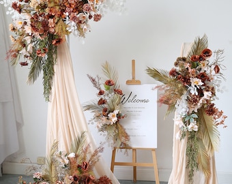 Pode incluir: Um cenário para uma cerimônia de casamento com dois arranjos florais com flores secas e folhagem. Os arranjos são colocados sobre tecido bege drapeado sobre suportes. Um cavalete de madeira exibe uma placa com a palavra "Bem-vindos" e uma mensagem de boas-vindas para o casamento.