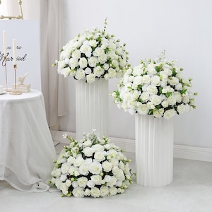 May include: Three white round floral arrangements with white roses and green leaves on white pedestal stands. The stands are fluted and have a classic design.