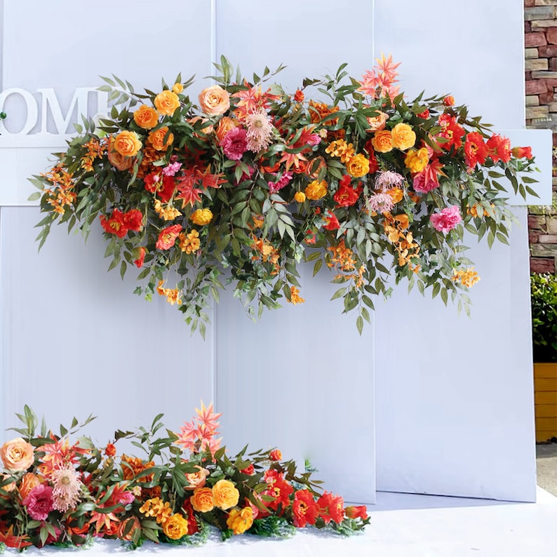 May include: A white backdrop with the word "HOME" in white letters, decorated with two garlands of artificial flowers. The garlands are made of orange, yellow, and pink flowers with green leaves.