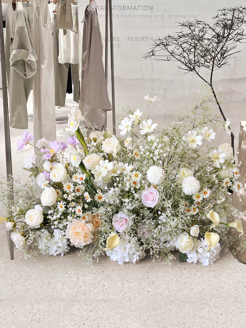May include: A large arrangement of white, pink, and peach flowers with green foliage. The flowers are arranged in a vase on a white surface. The text "Transformation" and "Pure Rebirth" is visible in the background.