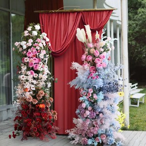 May include: Two tall floral arrangements, one with pink and white flowers and the other with blue and pink flowers, stand in front of a red curtain backdrop. The arrangements are made with artificial flowers and greenery.