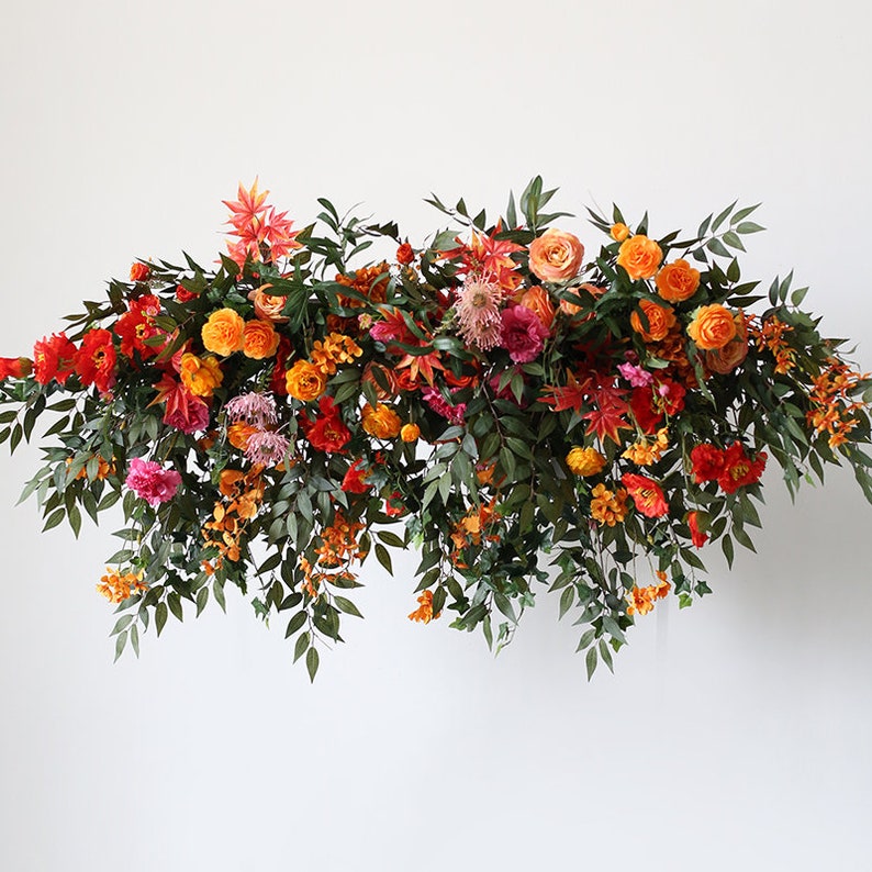 May include: A floral garland with a mix of orange, red, and pink flowers, and green leaves. The garland is hanging from a white wall.