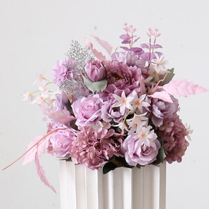 May include: A bouquet of artificial flowers in shades of purple and pink. The bouquet includes roses, hydrangeas, and other small flowers. The bouquet is arranged in a white vase with vertical grooves.