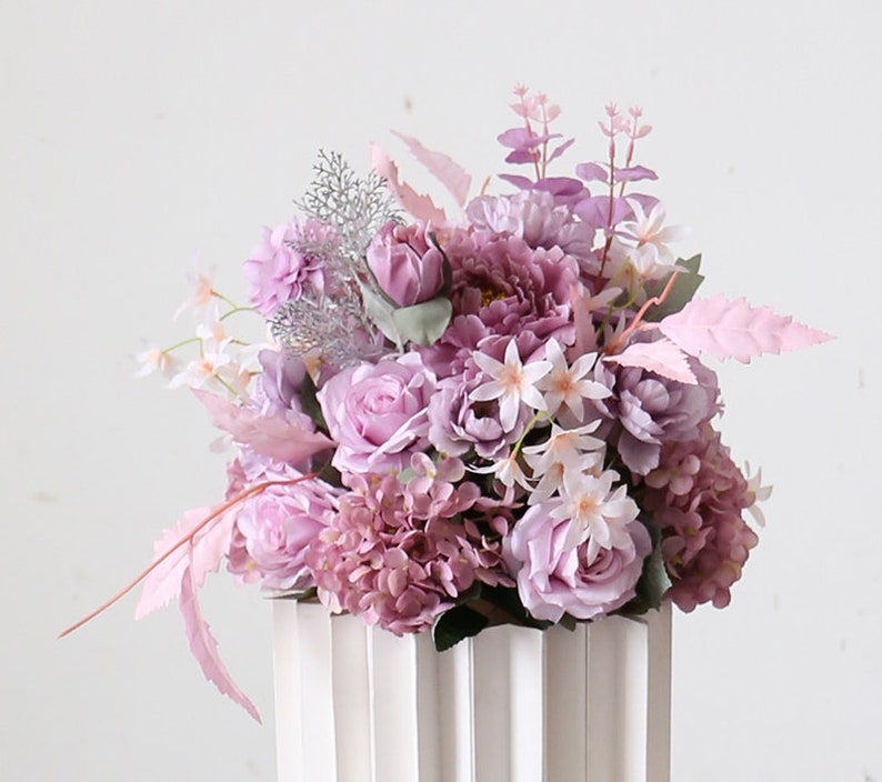 May include: A bouquet of artificial flowers in shades of purple and pink. The bouquet includes roses, hydrangeas, and other small flowers. The bouquet is arranged in a white vase with vertical grooves.