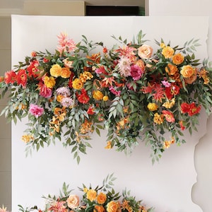 May include: A floral garland with orange, red, and pink flowers and green leaves. The garland is hanging on a white wall.