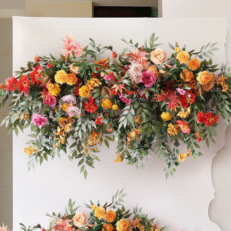 May include: A floral garland with orange, red, and pink flowers and green leaves. The garland is hanging on a white wall.