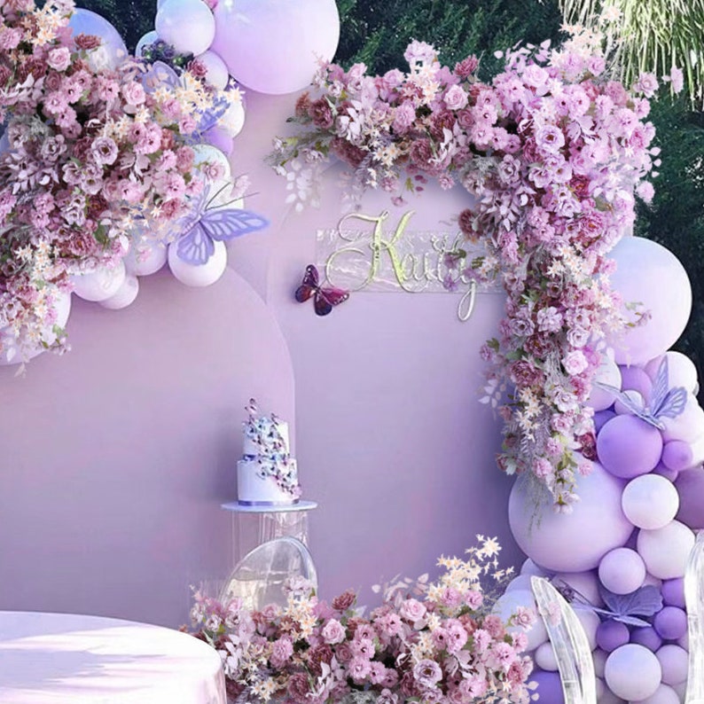 May include: A purple and white birthday party backdrop with a purple balloon arch, purple flowers, and a sign that says 'Keily'. The backdrop is decorated with butterflies and a cake on a clear stand.