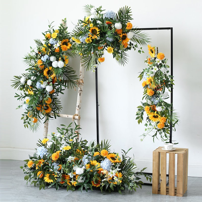 May include: A set of three artificial flower arrangements featuring sunflowers, white pom poms, and blue hydrangeas. The arrangements are displayed on a white wall with a black metal frame and a wooden ladder.