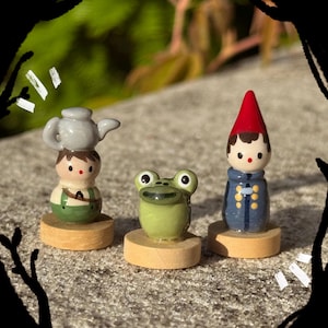 May include: Three small hand-painted figurines on circular wooden bases. One figure wears green overalls with a gray teapot on its head, the center figure is a green frog with large eyes, and the third figure wears a blue coat and a red pointed hat.