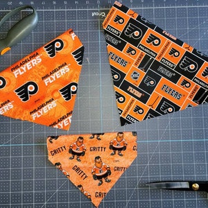 Può includere: Tre bandane di stoffa a tema Philadelphia Flyers. Una bandana presenta il logo dei Flyers e le parole "Philadelphia Flyers" ripetute in un motivo. Un'altra bandana presenta il logo dei Flyers e le parole "Flyers" ripetute in un motivo. La terza bandana presenta un personaggio dei cartoni animati chiamato Gritty, la mascotte dei Flyers, e la parola "Gritty" ripetuta in un motivo.