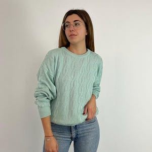 May include: A pale green cable knit jumper with a crew neck. The jumper has long sleeves and a textured pattern. The person is wearing blue jeans. The background is a plain white wall.