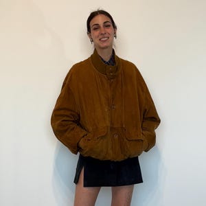 Op de afbeelding: Een bruine corduroy jas met knoopsluiting en twee voorzakken. De jas wordt gecombineerd met een zwarte rok en zwarte laarzen. Het model draagt roze sokken.
