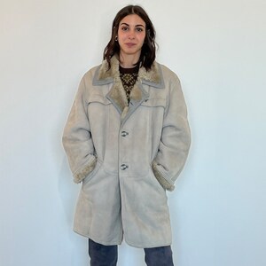 May include: A long, light beige shearling coat with a brown fur collar. The coat has a button closure and is worn by a person with dark hair and blue jeans.