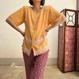 Op de afbeelding: Een vintage lichtoranje blouse met korte mouwen, versierd met bloemendetails en parelmoer knopen. De blouse heeft een kanten rand met franjes. Gecombineerd met een rode broek met bloemenprint.