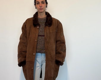 Puede incluir: Una chaqueta de ante marrón con cuello de piel sintética y cierre de botones. La chaqueta es larga y tiene un corte holgado. La modelo lleva pantalones vaqueros azules claros y botas marrones.