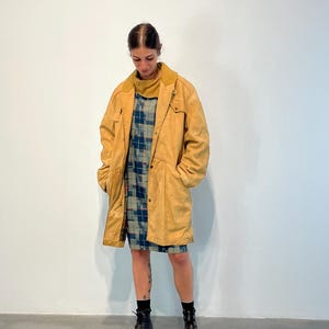 May include: A long, yellow suede coat with a button closure and pockets. The coat is worn over a plaid dress and black boots.