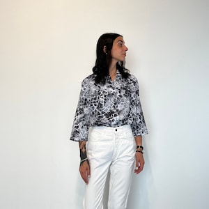 Può includere: Una persona indossa una camicia bianca con una stampa astratta in bianco e nero e jeans bianchi.