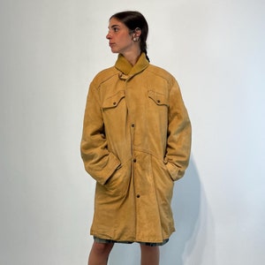 May include: A long, tan suede coat with a button-up front and a collar. The coat has two large pockets on the front and a belt at the waist.