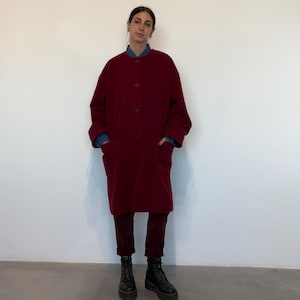Puede incluir: Un abrigo largo burdeos con bolsillos grandes y botones. El abrigo se lleva sobre una camisa vaquera y pantalones burdeos. Botas negras completan el conjunto. El abrigo es de color liso y parece estar hecho de un material suave.