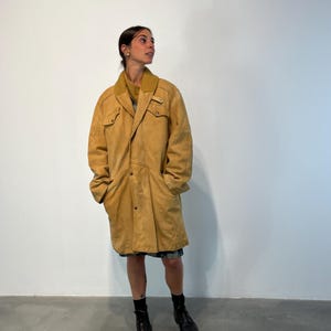 May include: A long, tan suede coat with a button closure and a collar. The coat has two large pockets on the front and a small pocket on the chest. The coat is worn by a person with dark hair and is standing on a concrete floor.