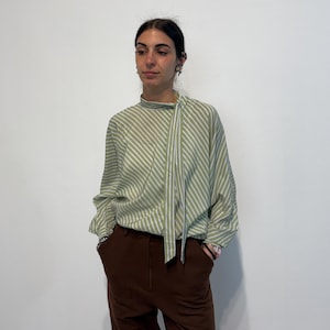 May include: A pale green and white striped blouse with a high neckline and a tie. The long-sleeved top is worn with brown trousers. The blouse has a relaxed fit and a subtle pattern, creating a classic and versatile look.