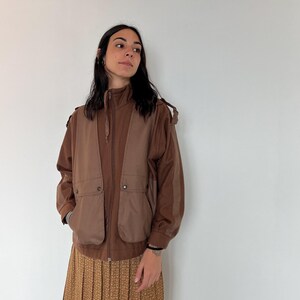 Puede incluir: Una chaqueta marrón con cierre de cremallera y un diseño tipo chaleco. La chaqueta tiene dos bolsillos en la parte delantera y una capucha con cordón.