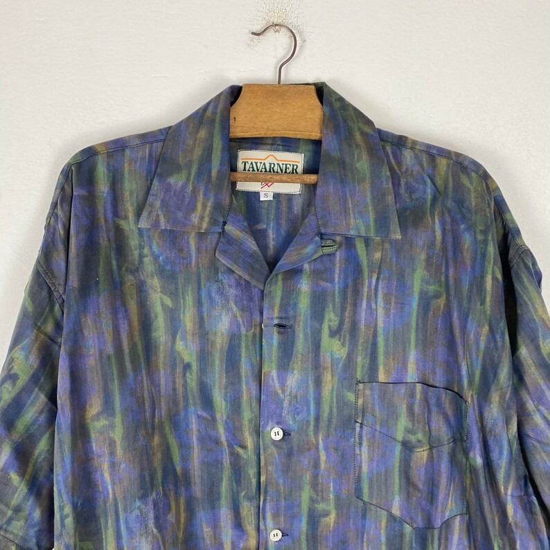 May include: A blue, green, and brown patterned short-sleeved button-up shirt with a collared neckline. The shirt has a pocket on the left side and a label that reads "Tavarner 92" in green and white.
