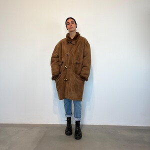 May include: A brown shearling coat with a fur collar and a toggle closure. The coat has two large pockets on the front and is worn over blue jeans.