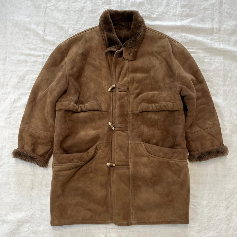 May include: A brown suede shearling coat with a fur collar and toggle closures. The coat has two large pockets on the front and a smaller pocket on the left side.
