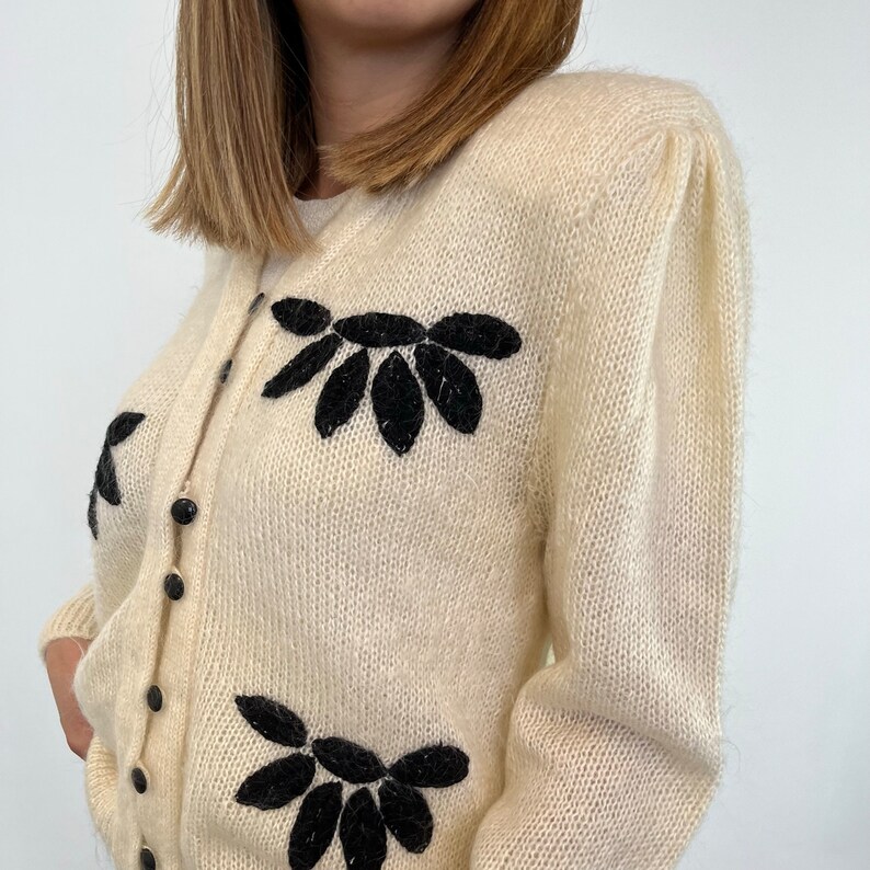 May include: A cream-coloured cardigan with black embroidered floral designs. The cardigan has a button-up front and long sleeves.