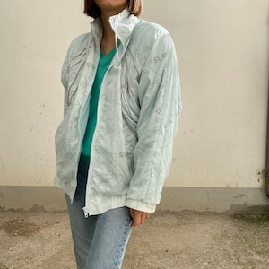May include: A light blue vintage jacket with a high collar and a zipper closure. The jacket has a subtle pattern and the word "Sun" is visible. The jacket is worn over a teal sweater and paired with blue jeans.