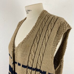 K&ouml;nnte beinhalten: Beige Strickweste mit marineblauen Streifen. Das &auml;rmellose Kleidungsst&uuml;ck hat einen V-Ausschnitt und wird mit Kn&ouml;pfen geschlossen. Das Strickmuster weist Zopfmusterdetails auf.