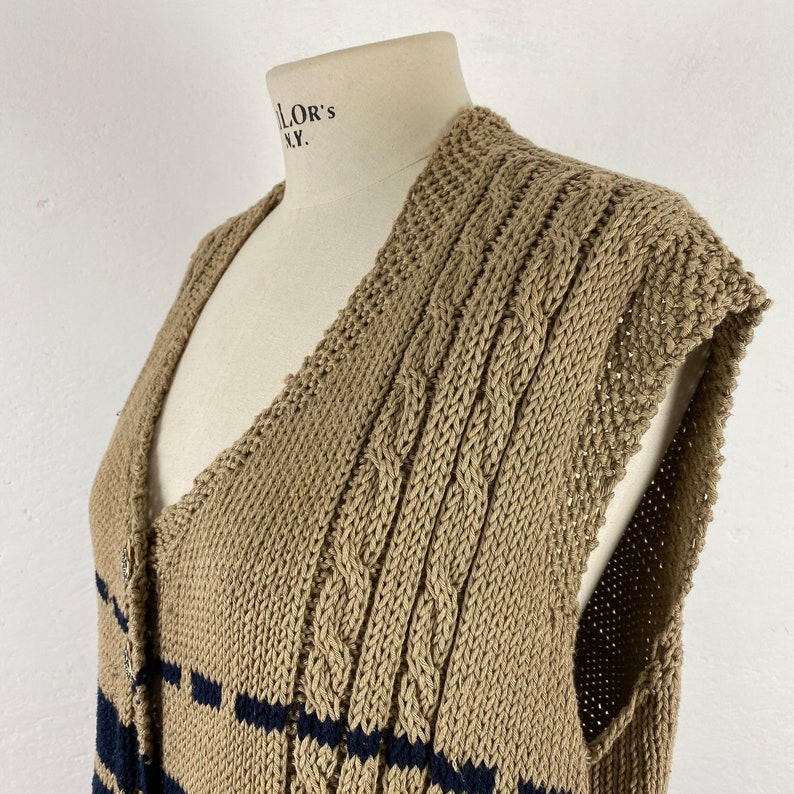 K&ouml;nnte beinhalten: Beige Strickweste mit marineblauen Streifen. Das &auml;rmellose Kleidungsst&uuml;ck hat einen V-Ausschnitt und wird mit Kn&ouml;pfen geschlossen. Das Strickmuster weist Zopfmusterdetails auf.