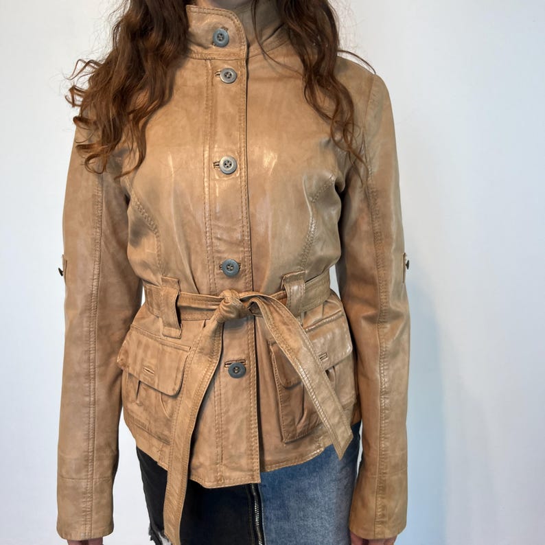 K&ouml;nnte beinhalten: Eine braune Lederjacke mit G&uuml;rtel und Taschen. Die Jacke hat einen Knopfschlie&szlig;e vorne und lange &Auml;rmel.