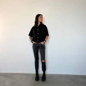 May include: A person wearing a black short-sleeved button-up shirt, black jeans with a knee rip, and black boots. The shirt has two chest pockets and is buttoned up. The jeans are a dark gray wash. The person is standing in front of a white wall.