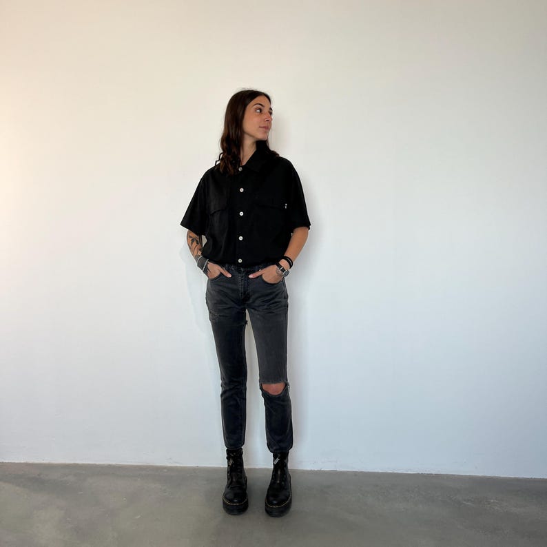 May include: A person wearing a black short-sleeved button-up shirt, black jeans with a knee rip, and black boots. The shirt has two chest pockets and is buttoned up. The jeans are a dark gray wash. The person is standing in front of a white wall.