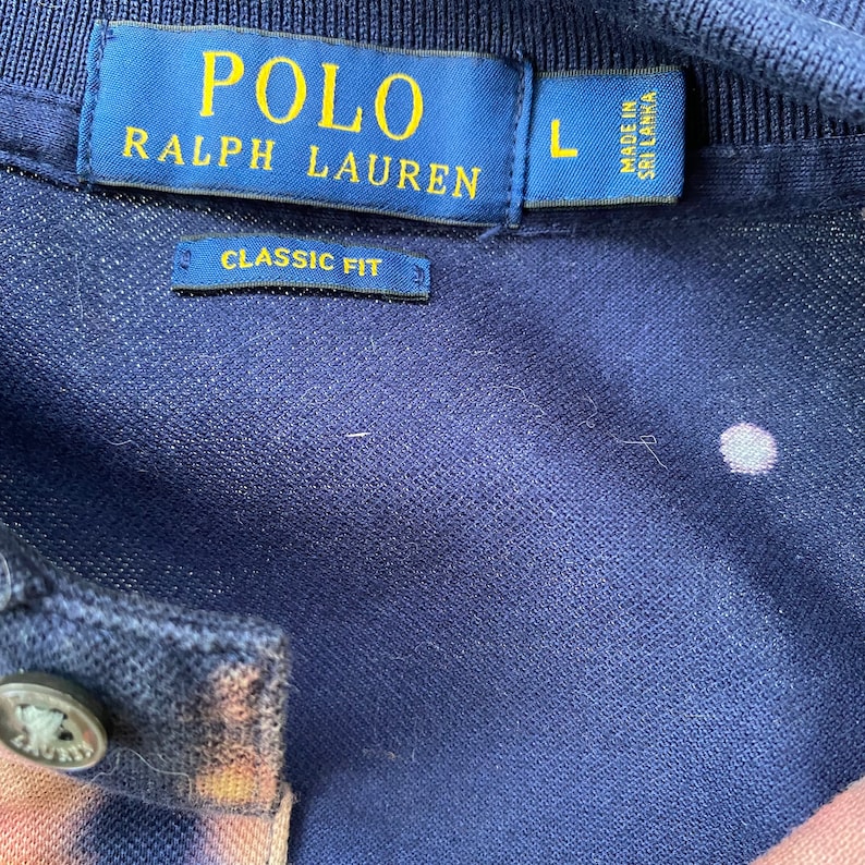 May include: A navy blue polo shirt with a white tag that reads "POLO RALPH LAUREN" and "CLASSIC FIT". The tag also indicates a size "L" and that the shirt was made in Sri Lanka.