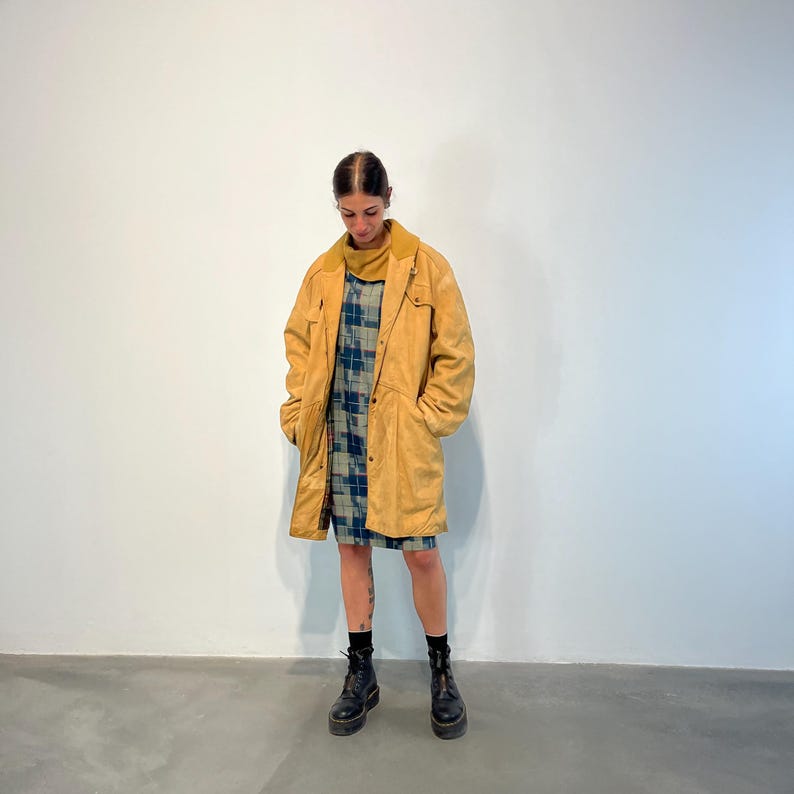 May include: A long, yellow suede coat with a button closure and pockets. The coat is worn over a plaid dress and black boots.