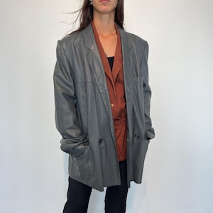 Op de afbeelding: Een grijs leren blazer met een dubbele rij knopen en een lange, ontspannen pasvorm.