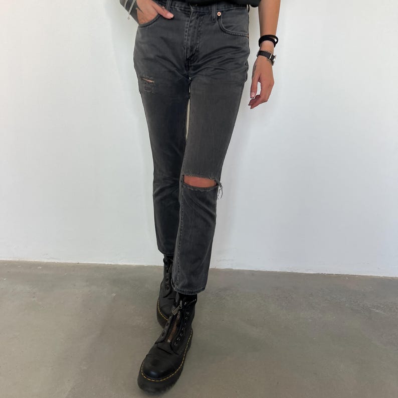 May include: Dark gray denim jeans with a distressed knee and a faded appearance. The jeans have a classic five-pocket design and are paired with black combat boots.