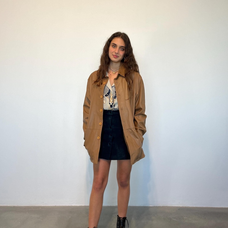 May include: A brown leather jacket with a button-up front and long sleeves. The jacket is worn over a white and blue patterned shirt and a black mini skirt.