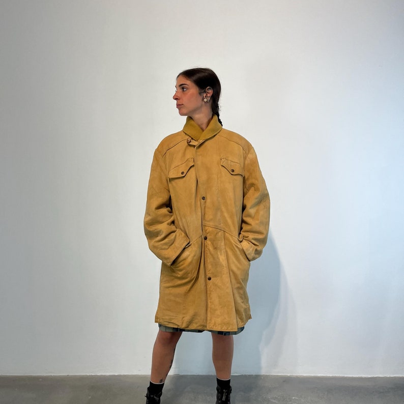 May include: A long, tan suede coat with a button-up front and a collar. The coat has two large pockets on the front and a belt at the waist.