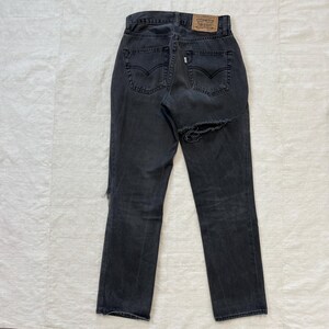 May include: Back view of a pair of faded black denim jeans with a distressed design. The jeans feature a high waist, straight leg, and a large tear on the right leg. A brown leather patch with a logo is visible at the waistband.
