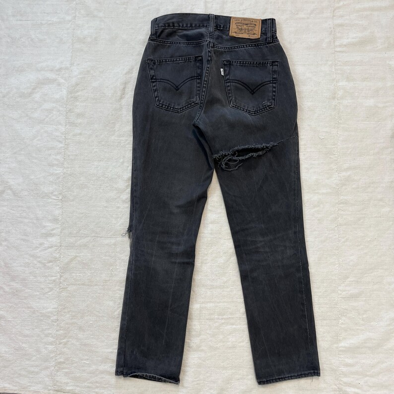 May include: Back view of a pair of faded black denim jeans with a distressed design. The jeans feature a high waist, straight leg, and a large tear on the right leg. A brown leather patch with a logo is visible at the waistband.