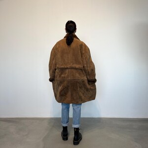May include: A brown suede and shearling coat with a belt. The coat is long and has a relaxed fit.