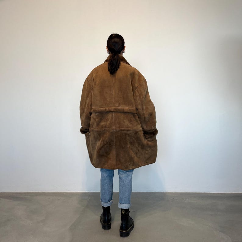 May include: A brown suede and shearling coat with a belt. The coat is long and has a relaxed fit.