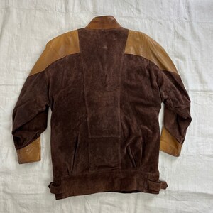 May include: A brown suede jacket with brown leather accents on the shoulders and sleeves. The jacket has a stand-up collar and a zipper closure.