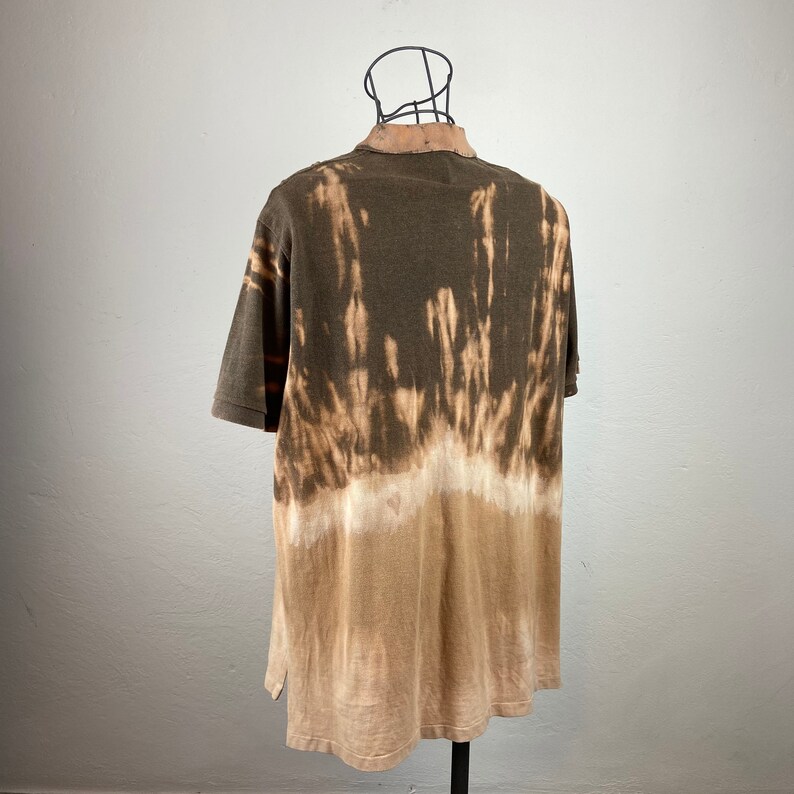 K&ouml;nnte beinhalten: Ein kurz&auml;rmeliges Poloshirt mit einem einzigartigen Batik-Effekt. Das Shirt wechselt von einem dunklen Olivgr&uuml;n oben zu einem hellen Beige unten, wobei gebleichte Streifen ein markantes Muster bilden. Der Kragen ist hellbraun.